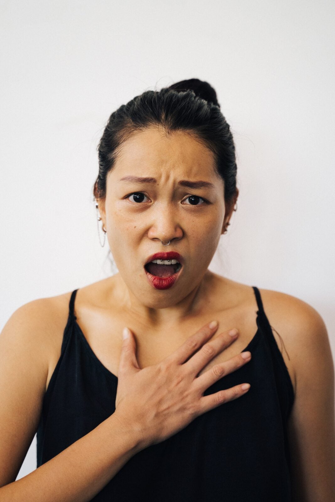 An Asian woman wearing a black tank top, holding her hand up to her chest with a shock look on her face. 