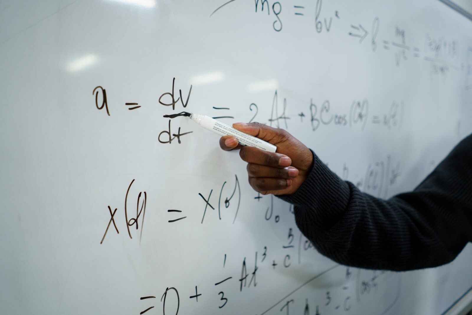 A man with dark skin, holding a marker and writing equations on a whiteboard.