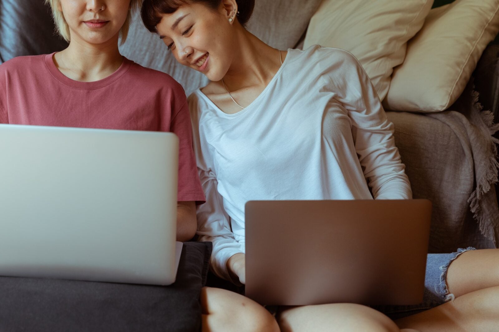 Two Asian women sitting on the floor in front of their sofa closely next to each other and using their laptops