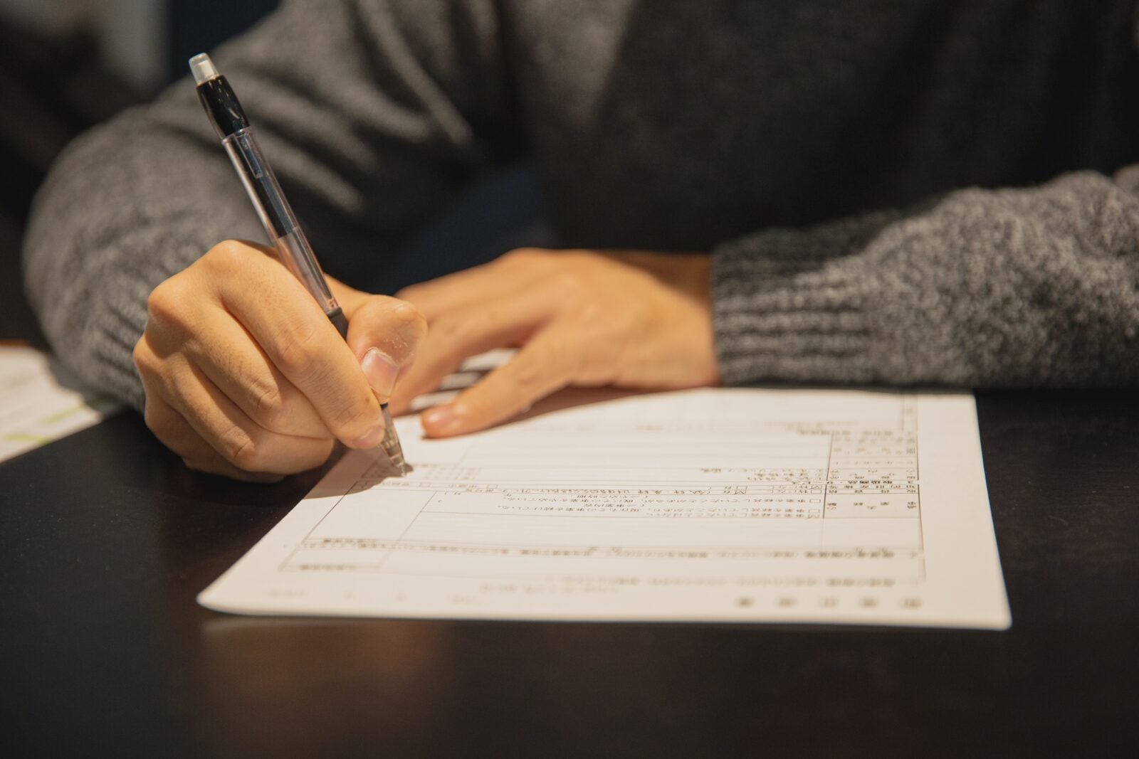 cropped of a person wearing a grey sweater, holding a pen and filling a form.