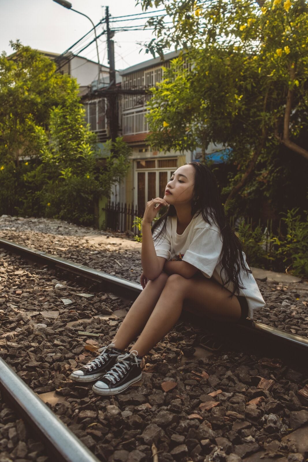 An Asian woman sitting on a train track railing with her head leaning on her hand. She is wearing shorts and a baggy shirt. 