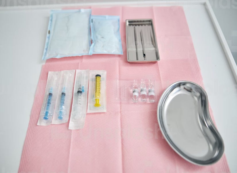 A Collection Of Medical Needles, Two Trays And A Vial Which Are Laid On Top Of A Pink Napkin]