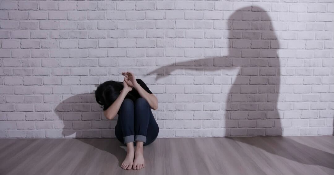 A Child Sitting On The Floor Against The Wall, Hiding Her Face With Her Arms While The Shadow Cast Along The Wall Is Of A Woman Pointing An Accusatory Finger At Her