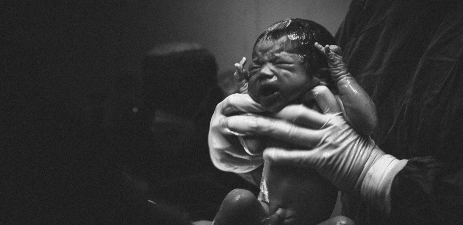 A Newborn Baby Held Up By Two Gloved Hands