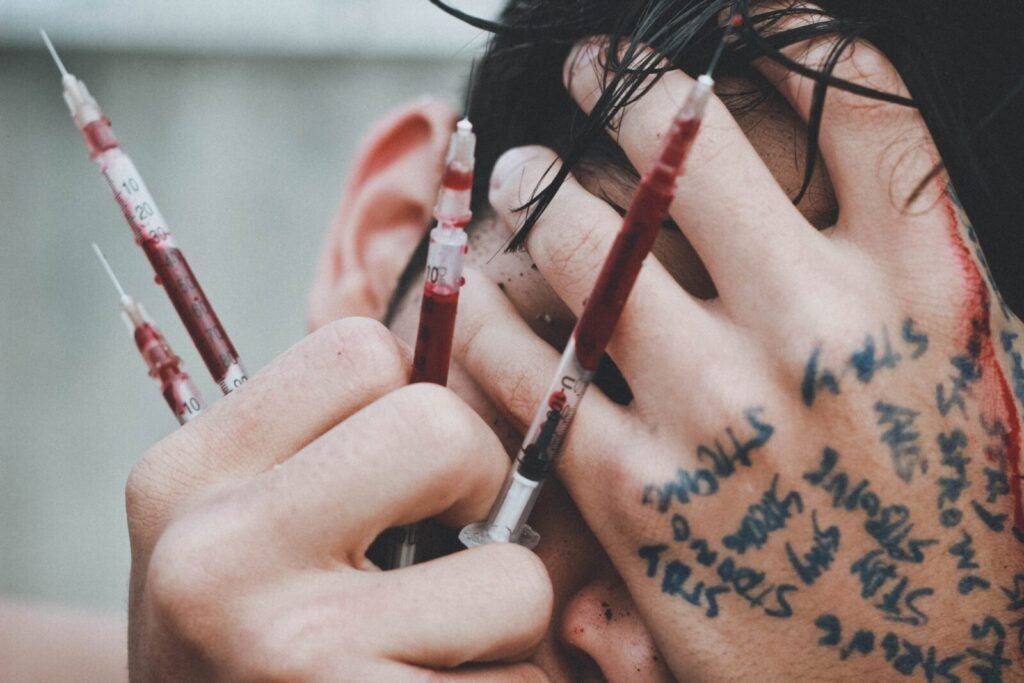 A Person Holding Multiple Needles Filled With Blood. Image Via Unsplash.